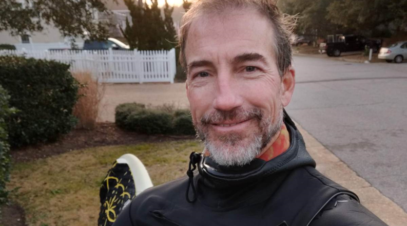 A man in a wet suit poses with a surfboard under his arm.