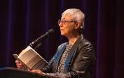 A woman reads from a book on a stage.