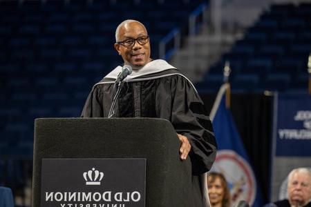 a man wearing academic regalia gives a speech standing at a podium 